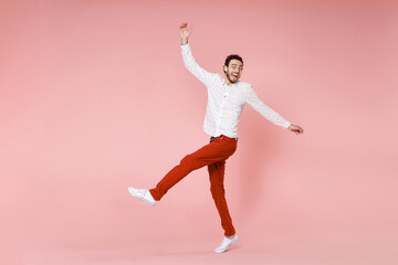 Wall Mural - Full length side view of excited young bearded man 20s wearing basic casual white shirt dancing spreading hands rising leg looking camera isolated on pastel pink color wall background studio portrait.