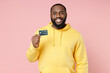 © ViDi Studio - Smiling cheerful young african american man 20s wearing casual yellow streetwear hoodie standing hold in hand credit bank card looking camera isolated on pastel pink color background studio portrait.