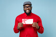 Smiling cheerful funny young bearded african american man 20s wearing casual red shirt eyeglasses cap standing hold gift certificate isolated on pastel blue color wall background studio portrait.