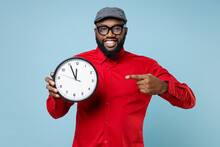 Smiling Young Bearded African American Man 20s Wearing Casual Red Shirt Eyeglasses Cap Standing Pointing Index Finger On Clock Looking Camera Isolated On Pastel Blue Color Background Studio Portrait.