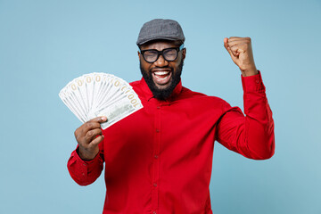 Canvas Print - Joyful young bearded african american man 20s in casual red shirt eyeglasses cap hold fan of cash money in dollar banknotes doing winner gesture isolated on pastel blue background studio portrait.