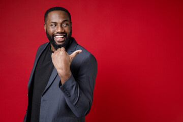 Wall Mural - Side view of cheerful funny young african american business man 20s wearing classic jacket suit standing pointing thumb aside on mock up copy space isolated on bright red background studio portrait.