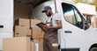 © VAKSMANV - Young handsome African American delivery man typing and tapping on tablet device standing near van on street with many ordered boxes. Male postman courier with shopping package. Shipment concept