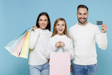 Wall Mural - Funny young parents mom dad with child kid daughter teen girl in sweaters hold package bags with purchases after shopping credit bank card isolated on blue background. Family day parenthood concept.