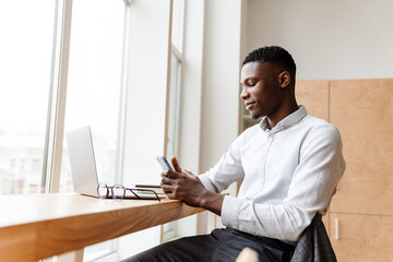 Poster - Afro american pleased man using mobile phone while working with laptop