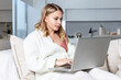 © Анна Демидова - Young blond woman is sitting on a sofa with a laptop in a large bright living room. Remote work and distance education.
