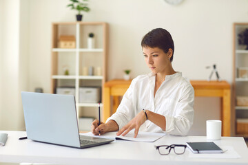 Wall Mural - Young woman looking at laptop and making notes during online lesson