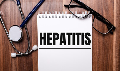 The word HEPATITIS is written on white paper on a wooden background near a stethoscope and black-framed glasses. Medical concept