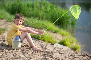 A little boy with a fishing net and a jar sits on the banks of a river or lake.