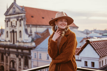 Fashionable happy smiling woman wearing trendy beige hat, rhinestones earrings, brown faux fur coat, posing on balcony with beautiful view on European city. Copy, empty space for text
