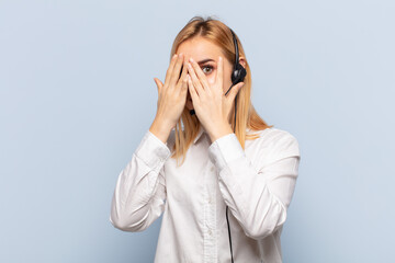 Wall Mural - young blonde woman covering face with hands, peeking between fingers with surprised expression and looking to the side