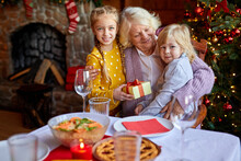Senior Woman Sitting At A Table With Grandchildren, Having Fun At Christmas Dinner, Enjoy Time Together, Sit By Nicely Decorated Christmas Tree