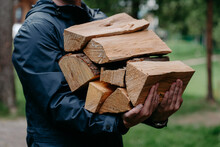 Cropped Shot Of Faceless Man In Black Jacket Carries Pile Of Firewood Poses Against Blurred Forest Background.