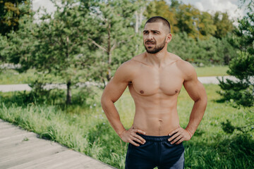 Horizontal shot of thoughtful shirtless sportsman keeps hands on waist, looks away demonstrates his muscular body, poses over nature background. Active bodybuilder goes in for sport, has daily workout