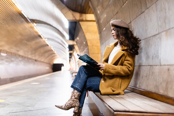 stylish woman in beret and autumn clothes reading magazine on metro platform