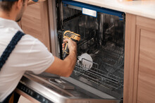 A Man Or A Service Worker In Special Clothing Uses A Screwdriver To Fasten To Neighboring Cabinets Or Dismantle The Dishwasher.