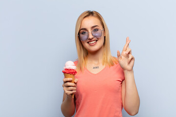 Wall Mural - young blonde woman smiling and anxiously crossing both fingers, feeling worried and wishing or hoping for good luck