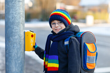 Little school kid boy of elementary class walking to school during snowfall. Happy healthy child with glasses pushing button for traffic lights. With backpack or satchel in colorful winter clothes.