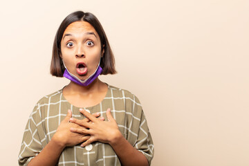 Canvas Print - young hispanic woman feeling shocked and surprised, smiling, taking hand to heart, happy to be the one or showing gratitude