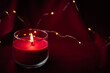 A red aroma candle burns and stands on red background with lights from garlands. Christmas mood