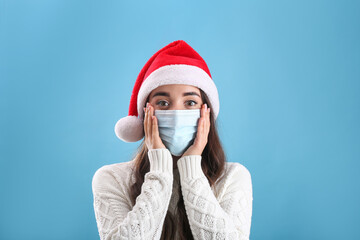 Wall Mural - Beautiful emotional woman wearing Santa Claus hat and medical mask on light blue background