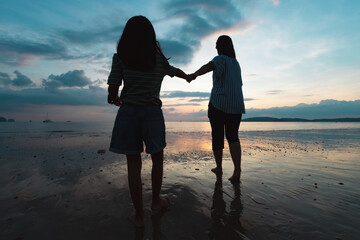 Silhouette of asian mother and daughter holding hand and walking on the beach together at the sunset time with beautiful sea and sky. Family enjoy with nature concept.