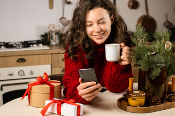 Happy young woman in red sweater making video call with smartphone, holding cup of coffee, sitting at kitchen table.Sharing data on social media,chatting with friend.Christmas celebration in distance