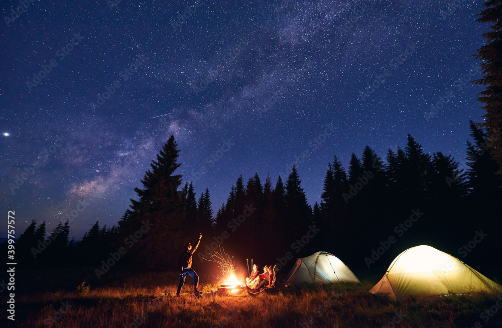 Man showing his friends Milky Way over tent city. Guys are sitting by the campfire on the background of the spruce forest and enjoying the evening starry sky. Outdoor recreation, touristm concept - obrazy, fototapety, plakaty 