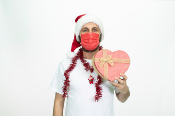 Wall Mural - Man wearing a Santa hat. He has a red corona mask on his face. He is holding a gift box in the shape of a heart.