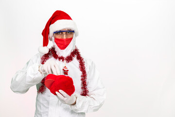 Wall Mural - Health worker wearing protective clothing. He wears a santa hat on his head. He has a red corona mask on his face. He is holding a gift box in the shape of a heart.