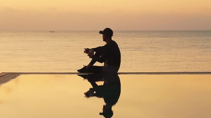 Wall Mural - Silhouette of a young man in sportswear and a cap sitting on the edge of the pool by the sea with a thermo mug in his hands, drinks coffee and enjoying the sunrise