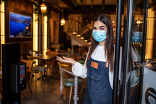 Portrait Of A Young Caucasian Female Waitress Wearing An Apron, Face Mask And Gloves, Standing At The Entrance Of A Drink Establishment And Welcoming Customers Of The Store.