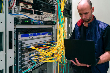 The system administrator with a laptop is in the server room. A man sets up a central router in a datacenter. Configuring server hardware.