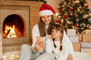 Christmas online, family congratulate someone with holiday, smiling european mother and daughter using mobile phone for video call, sitting on floor near fireplace and decorated fir tree.