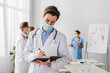 © LIGHTFIELD STUDIOS - Doctor in medical mask writing in notebook with blurred colleagues working on background
