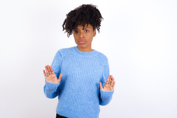 Wall Mural - Serious Young beautiful African American woman wearing blue knitted sweater against white wall pulls palms towards camera, makes stop gesture, asks to control your emotions and not be nervous