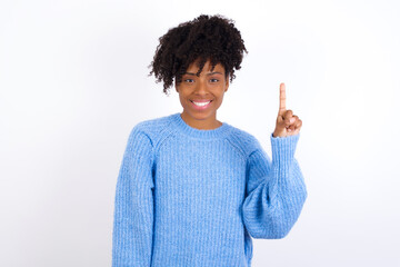 Wall Mural - Young beautiful African American woman wearing blue knitted sweater against white wall showing and pointing up with finger number one while smiling confident and happy.