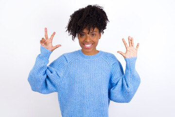 Wall Mural - Young beautiful African American woman wearing blue knitted sweater against white wall showing and pointing up with fingers number eight while smiling confident and happy.