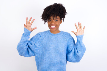 Wall Mural - Young beautiful African American woman wearing blue knitted sweater against white wall showing and pointing up with fingers number nine while smiling confident and happy.