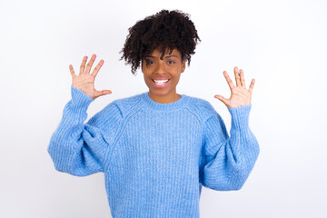 Wall Mural - Young beautiful African American woman wearing blue knitted sweater against white wall, showing and pointing up with fingers number ten while smiling confident and happy.