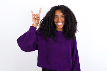 Wall Mural - Young beautiful African American woman wearing knitted sweater against white wall doing a rock gesture and smiling to the camera. Ready to go to her favorite band concert.