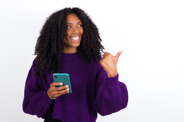 Wall Mural - Young beautiful African American woman wearing knitted sweater against white wall using and texting with smartphone pointing and showing with thumb up to the side with happy face smiling