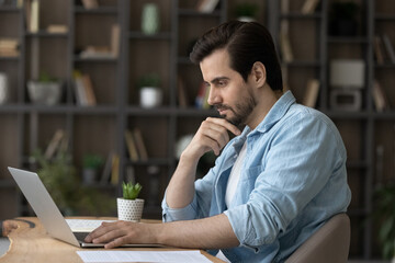 Wall Mural - Pensive millennial Caucasian man sit at desk at home office look at laptop screen working online with documents. Thoughtful young male employee use computer thinking analyzing project on web.