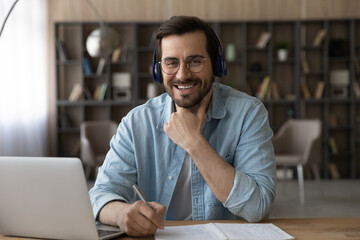 Portrait of smiling millennial male student in headphones sit at desk at home study online on laptop. Happy young Caucasian man in earphones work distant with client or customer on computer.