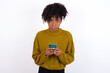 © Jihan - Portrait of a confused Young beautiful African American woman wearing knitted sweater against white wall holding mobile phone and shrugging shoulders and frowning face.