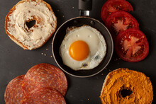 A Flat Lay Breakfast Or Snack On Black Wood For One Person: A Sesame Bagel Toasted And Sliced In Half With  Cream Cheese On, Slices Of Tomat,  Sunny Side Up Fried Egg And Artisan Salami On Black Wood