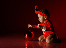 Curious Infant Baby Girl Toddler In Red Bodysuit And Headband With Bow Plays With Catches Rolling Christmas New Year Fir Tree Decoration Big Ball Over Dark Background. Translation: Happy New Year