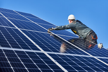 Worker standing on ladder and installing photovoltaic solar panel system. Male electrician in white safety helmet under blue sky. Concept of alternative energy and power sustainable resources.