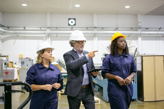 Serious engineer and diverse female factory workers in hardhats walking on plant floor and talking, man pointing at machines. Front view. Industrial occupation concept