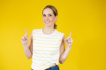 Wall Mural - Portrait of a happy young woman dressed in casual t-shirt pointing fingers up at copy space isolated over yellow background